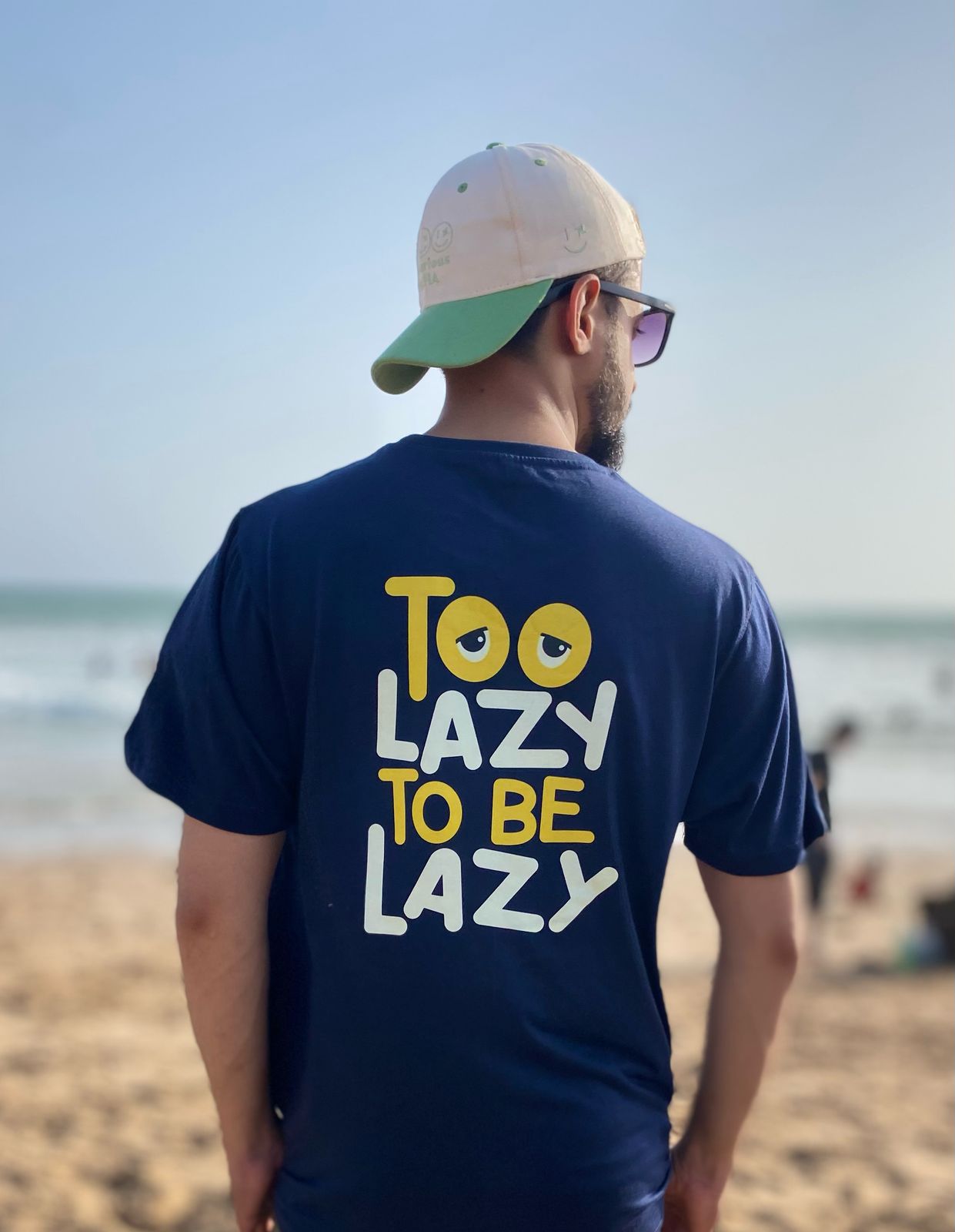 Man wearing a blue t-shirt with 'Too Lazy To Be Lazy' text on a beach