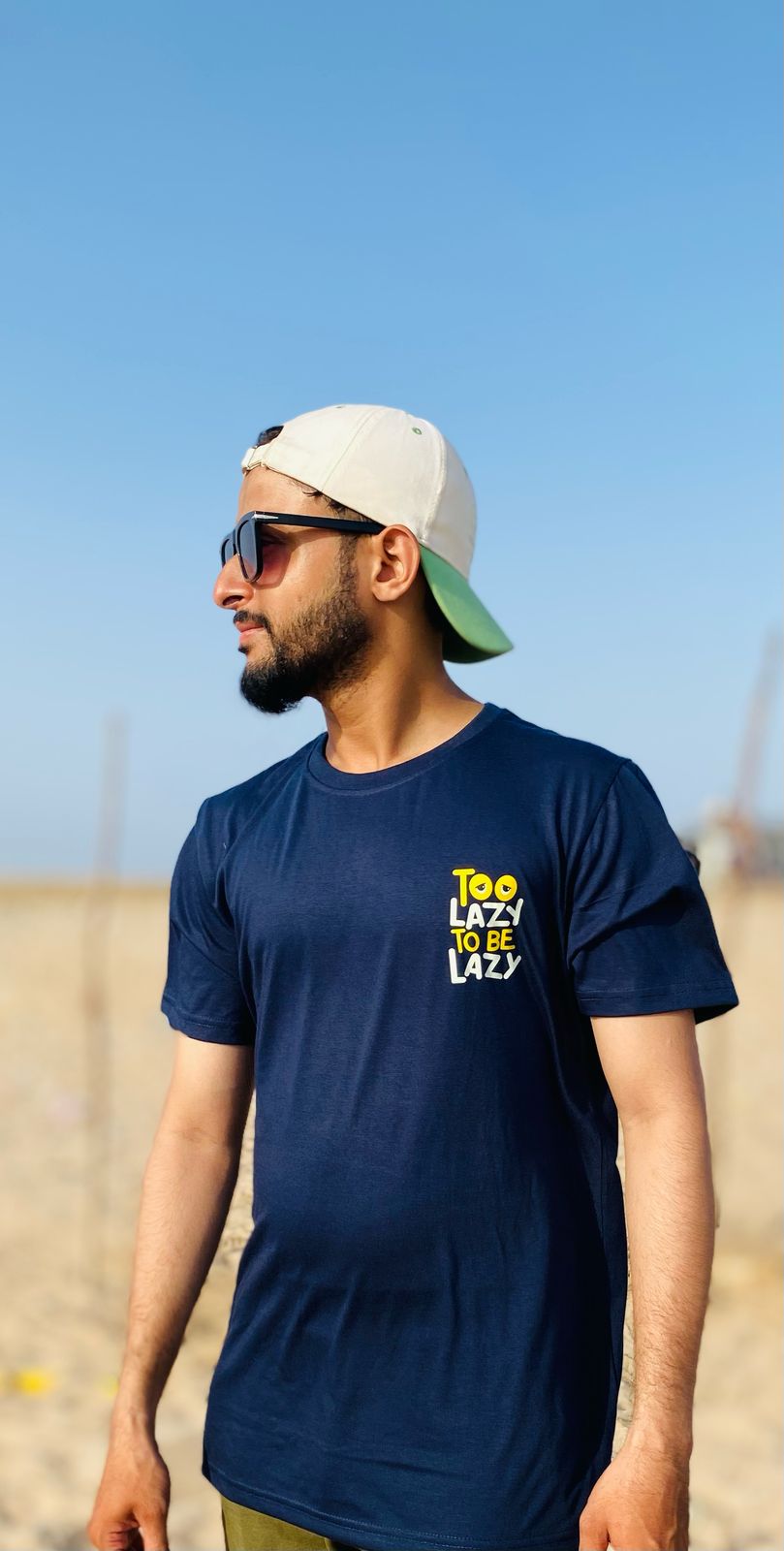 Man wearing a navy blue t-shirt with 'Too Lazy' text, sunglasses, and cap outdoors.