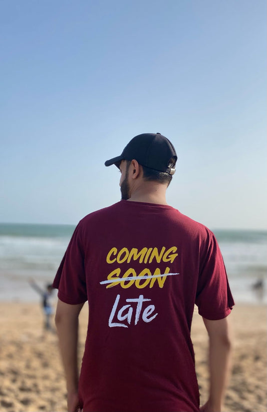 Person wearing a maroon t-shirt with 'COMING SOON Late' text on a beach